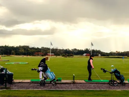 Kvinnor tränar golf på en driving range i Sverige. Solstrålar bryter igenom molnen.