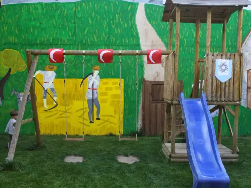 Un niño observa un columpio y un tobogán azul, parte de unos juegos medievales para niños, con un mural de fondo.