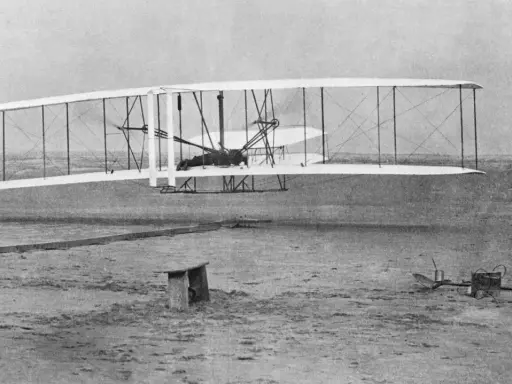 Wright Flyer mit Doppelruder hebt ab, Pilot liegt auf dem Flügel. Ein Mann beobachtet den historischen Flug.