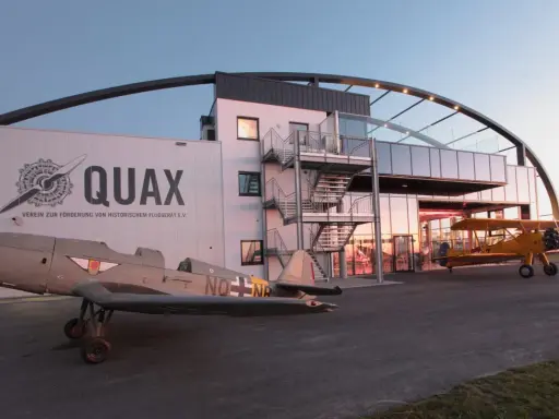 Zwei historische Flugzeuge stehen vor dem Quax Hangar. Ein graues Propellerflugzeug im Vordergrund, ein gelbes Doppeldeckerflugzeug daneben.