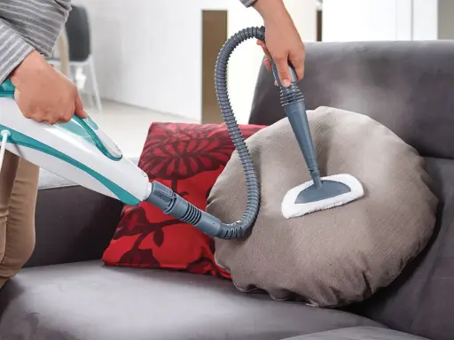 Persona pulisce un divano non sfoderabile con vaporetto, igienizzando i cuscini con vapore.