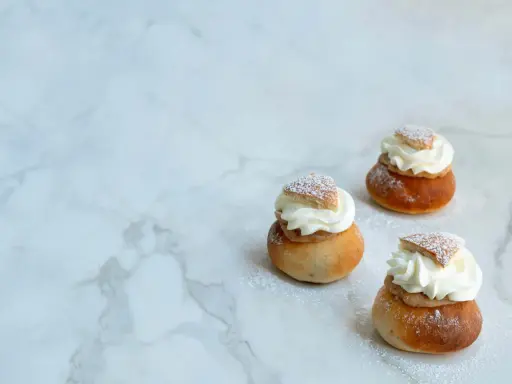 Tre semlor utan mandelmassa, toppade med vispgrädde och pudersocker, ligger på ett marmorbord.