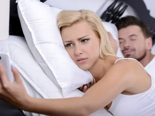 Donna bionda a letto con smartphone, sguardo preoccupato. L'uomo dorme accanto, forse ignaro del tradimento.
