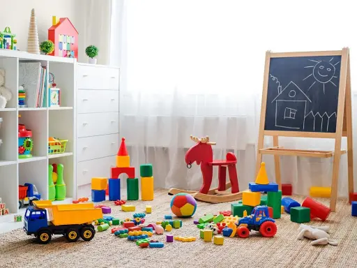 Kolorowy pokój zabaw dla dzieci w domu. Na dywanie rozrzucone klocki, ciężarówka, traktorek, huśtawka-łoś i tablica z rysunkiem domku.