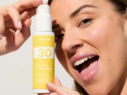 Une femme souriante tient un flacon de crème solaire visage SPF 30. La quantité de crème solaire est parfaite pour une protection optimale.