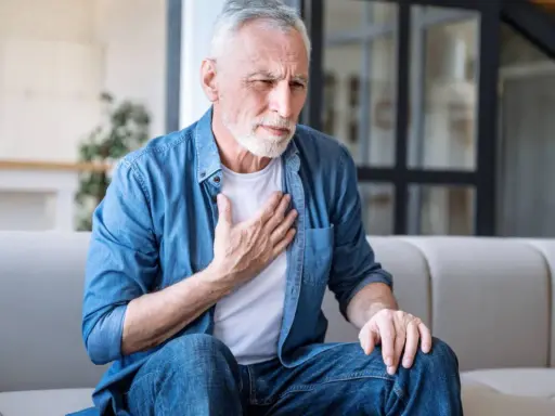 Älterer Mann mit COPD hält sich die Brust. Er sitzt auf einem Sofa und sieht besorgt aus.