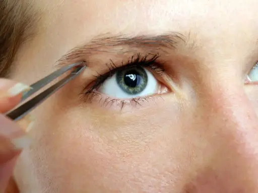 Une main utilise une pince à épiler pour affiner des sourcils, créant une ligne élégante au-dessus d'un œil bleu-vert.