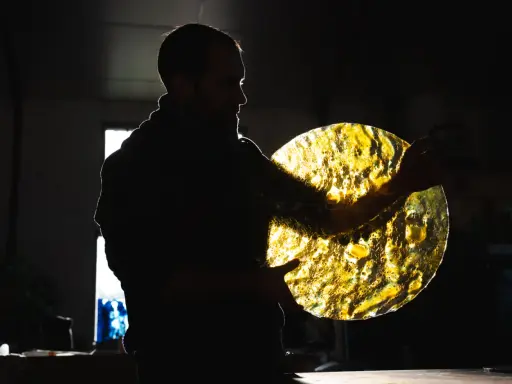 Artisan tenant un disque de verre texturé, illuminé par derrière. Les reflets dorés rappellent des vitraux.