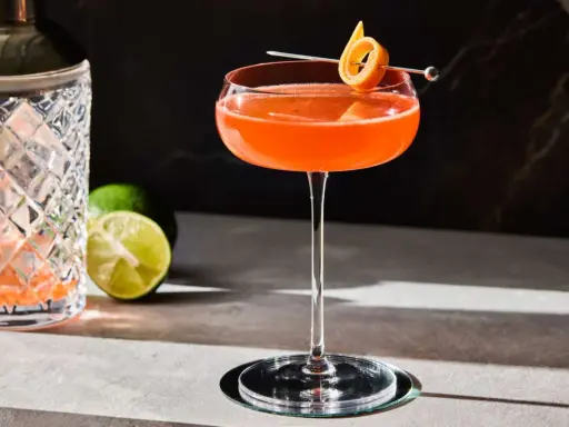 En vackert orangefärgad Division Bell cocktail med en apelsinzest som garnityr, bredvid en cocktailshaker och limeklyftor.