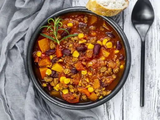 Herzhaftes Chili sin Carne mit Bohnen, Mais und Kürbis, garniert mit Rosmarin. Dazu ein Stück Brot und ein Löffel.