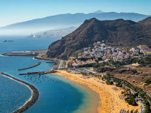 Bunte Häuser schmiegen sich an steile Hänge, während ein goldener Strand und das blaue Meer die Küste von Spanien zieren. Im Hintergrund erheben sich majestätische Berge.