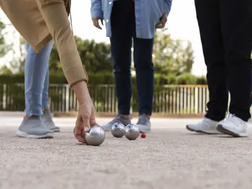 Une main de nom joueur de pétanque français saisit une boule argentée sur un terrain de gravier. D'autres boules et un cochonnet sont visibles.