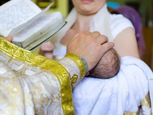 Ksiądz w złotej szacie trzyma dziecko do chrztu, czytając z księgi.