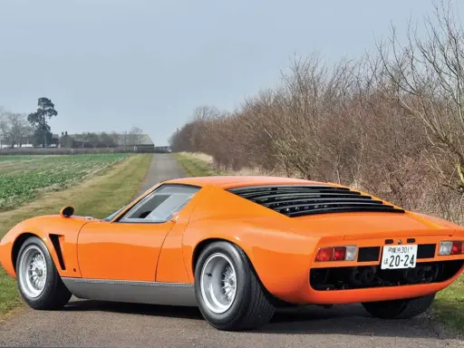 Ein leuchtend oranger Lamborghini Jota parkt auf einer einsamen Landstraße, umgeben von Feldern und kahlen Bäumen.