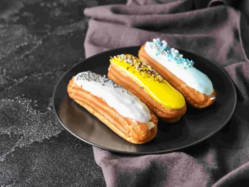 Trois éclairs gourmands sur une assiette noire : un blanc pailleté, un jaune croquant, un bleu ciel festif. La pâte à choux éclair inratable est prête à être dégustée.