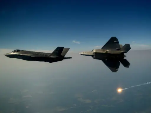 Zwei Tarnkappen-Kampfflugzeuge, ein F-35 und ein F-22, fliegen im Tandem über den Wolken. Ein F-22 schießt eine Leuchtrakete ab.