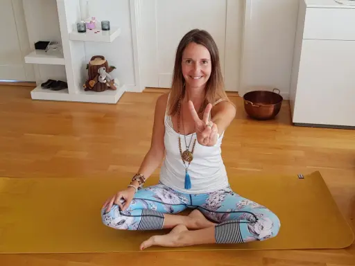 En kvinna sitter i lotusställning på en gul yogamatta, redo för morgon meditation. Hon ler och gör en fredstecken.