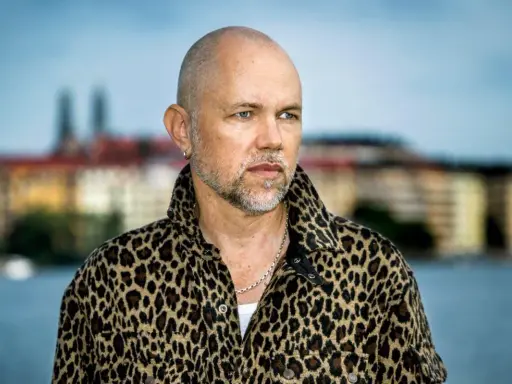 Peter LeMarc i leopardmönstrad jacka, redo för en konsert. Bakgrunden visar en stad vid vattnet.