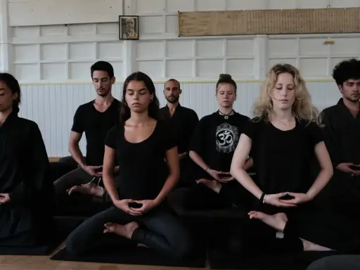 En grupp människor sitter i meditation, klädda i svarta kläder. En zen master leder sessionen.