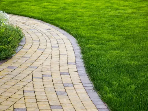 Vialetto ingresso casa curvo, pavimentato con blocchi di pietra chiara e scura, fiancheggiato da prato verde brillante e vegetazione rigogliosa.