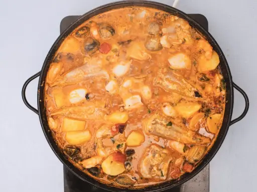 Un suquet de peix burbujeante en una paellera, con trozos de pescado, patatas, gambas y mejillones en una salsa naranja.