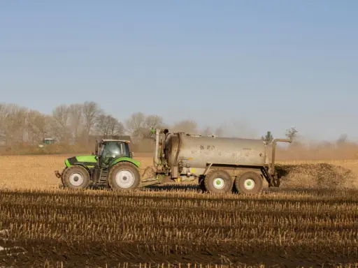 Zielony traktor z przyczepą rozprowadza obornik bydlęcy na polu.