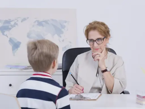 Chłopiec rozmawia z nauczycielką o ocenach. Nauczycielka słucha uważnie, notując coś na kartce.