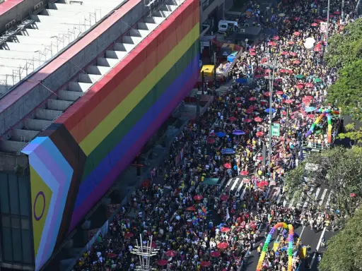 A massive crowd fills the street for what appears to be the biggest pride parade, with a giant rainbow flag draped over a building.