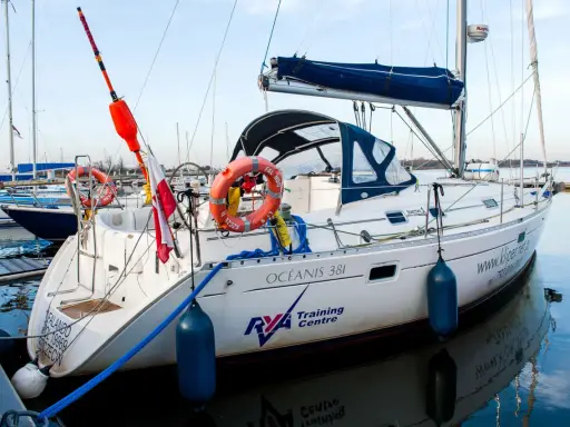 Biały jacht "Océanis 381" z czerwonymi kołami ratunkowymi i polską flagą, cumujący w marinie. To jakby patent na jacht, gotowy do rejsu.