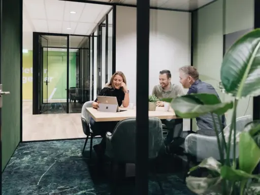Tre kollegor skrattar vid ett mötesbord i en modern, inredd verkstad. En kvinna visar något på sin laptop.