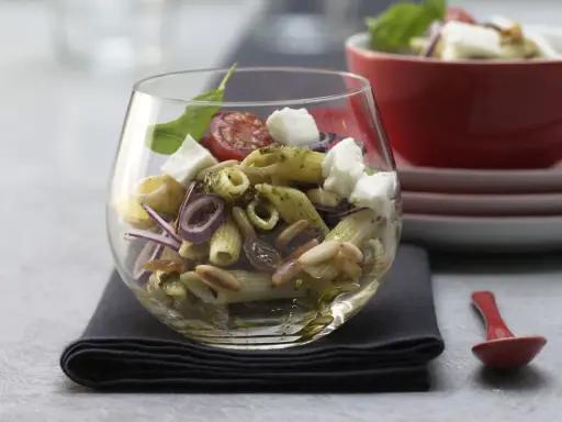Ein köstlicher Nudel Pesto Salat mit Mozzarella, Pinienkernen, Rosinen und roten Zwiebeln in einem Glas serviert.