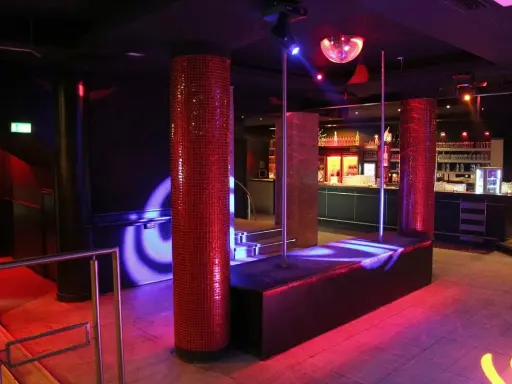 Interno di una boite de nuit con luci soffuse, un palo da lap dance, un bancone bar e una sfera da discoteca.