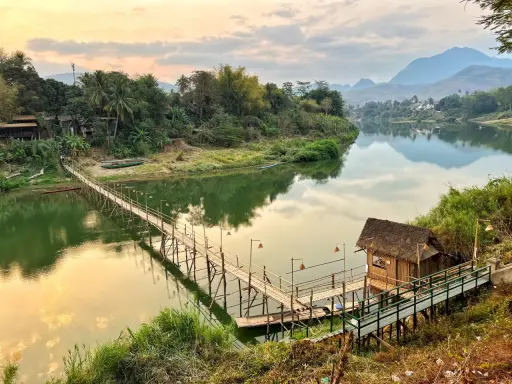 Drewniany mostek nad rzeką, bambusowa chatka i góry w tle. To jedne z wielu **laos atrakcje** warte zobaczenia.