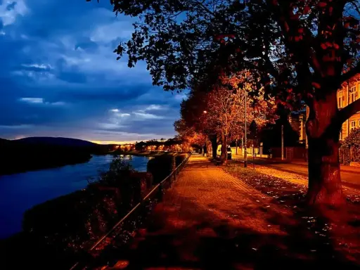 Un sentiero illuminato da lampioni lungo un fiume, con alberi dalle foglie arancioni e rosse. Una magica sera d'ottobre sui pascoli.