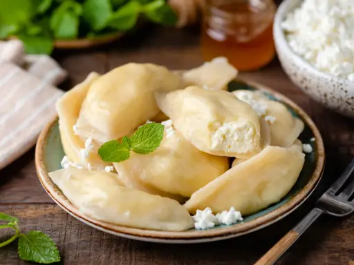 Pyszne pierogi z serem, udekorowane miętą i posypane białym serem. Obok słoiczek miodu i miseczka z serem.