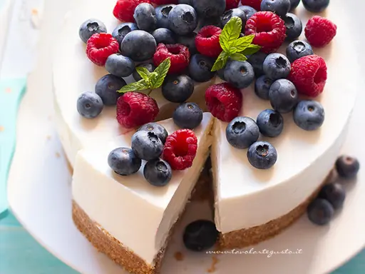 Cheesecake allo yogurt fresca e cremosa, decorata con lamponi e mirtilli succosi. Una fetta è già stata tagliata, invitando a gustarla.