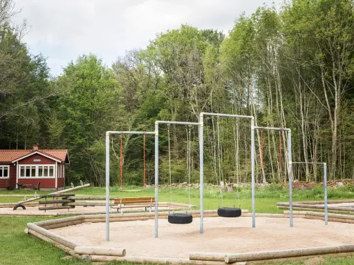 Lekplats med gungor och sandlåda vid en röd stuga, en idyllisk plats som påminner om nyrups naturhotell.