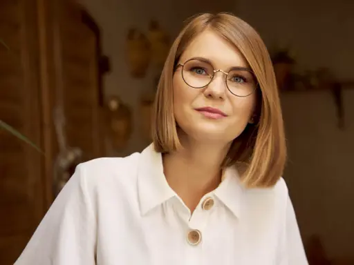 Kobieta w białej koszuli z guzikami, nosząca okulary w cienkich oprawkach, patrzy wprost.