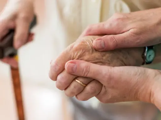 Dwie dłonie, jedna starsza, druga młodsza, trzymają się nawzajem. Laska w tle sugeruje trudności z poruszaniem się, być może związane z chorobą Parkinsona i jej dziedziczeniem.