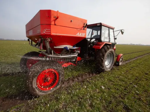 Czerwony traktor z rozsiewaczem nawozu na wiosennym polu. Odpowiedni nawóz pod warzywa na wiosnę to klucz do obfitych plonów.