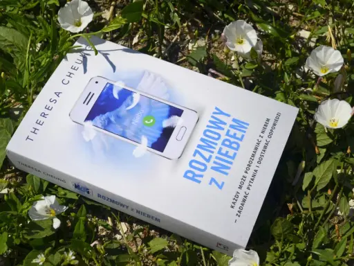 Książka "Rozmowy z niebem" leży wśród zieleni i białych kwiatów. Na okładce telefon z napisem "Niebo" i piórami.