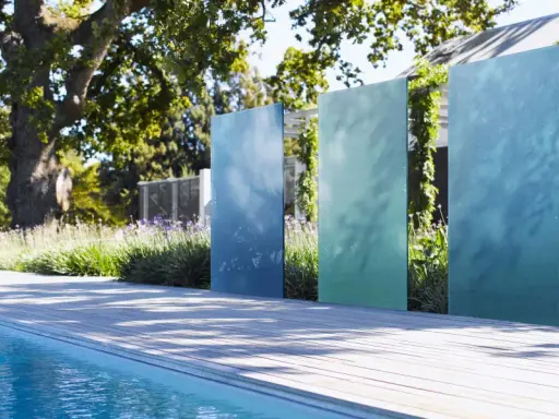 Ausgefallener Sichtschutz Garten selber machen: Vertikale, blaue und grüne Metallplatten vor üppiger Bepflanzung und Pool.