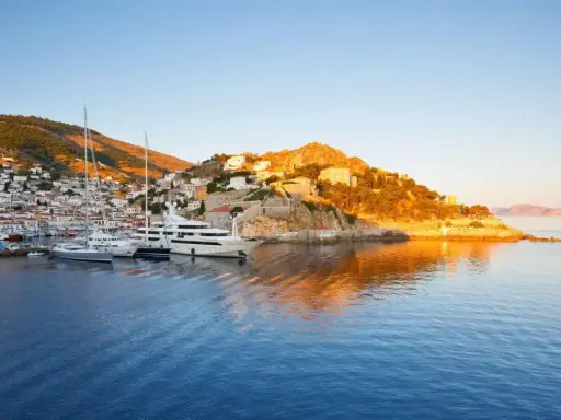 Solnedgång över hamnen på Hydra, Grekland. Lyxyachter guppar i det blå vattnet framför en vit stad som klättrar uppför en grönskande kulle.