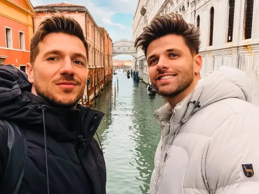 Two men enjoying a romantic gay Venice trip, smiling for a selfie with canals and historic buildings behind them.