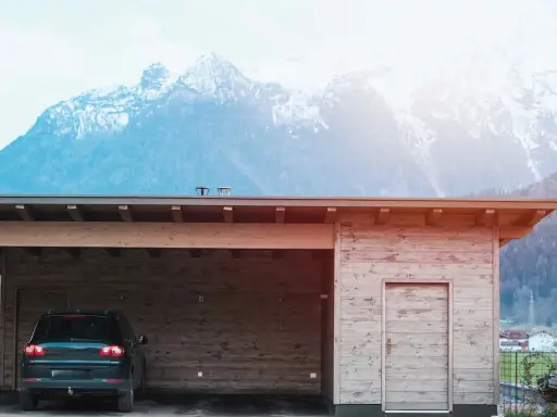 En nybyggd carport i trä med en bil parkerad under. Bakom syns snöklädda berg.