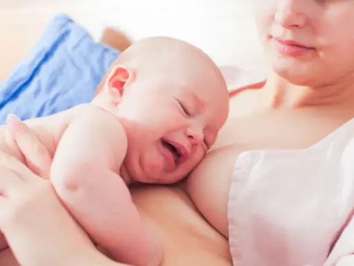 Ein Baby schreit beim Stillen, die Augen geschlossen, das Gesicht verzogen. Die Mutter hält es sanft.