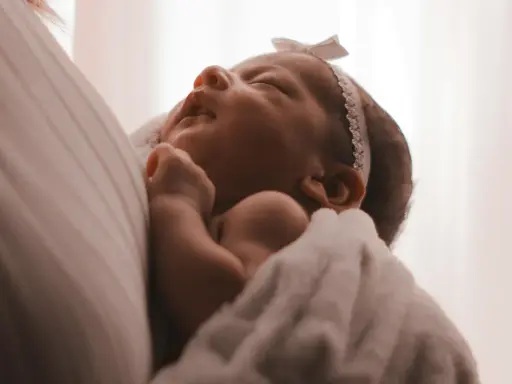Un neonato avvolto in una coperta bianca, con un fiocco rosa in testa. I suoi piccoli tremori sono quasi impercettibili mentre dorme serenamente.