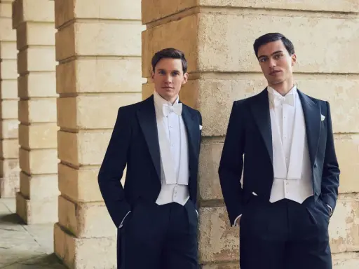 Two men in formal attire, a white tie event, stand by a stone colonnade.