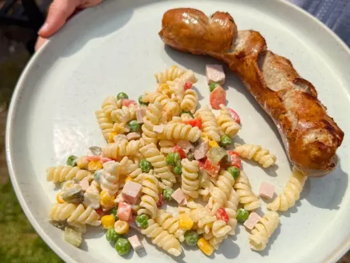 Gegrillte Wurst mit einem cremigen Nudelsalat als Beilage, garniert mit Erbsen, Mais und gewürfeltem Schinken.