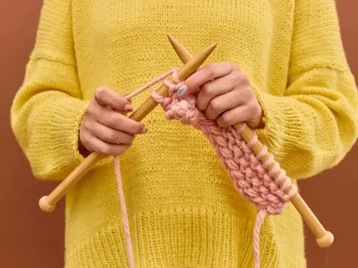 Manos tejiendo un suéter amarillo con hilo rosa, mostrando cómo hacer ochos de 4 puntos.